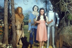 Judy Garland as Dorothy, a girl in a pink dress stands on a yellow brick road with Wizard of Oz wax figures at Movieland—a dog, lion, scarecrow, and tin man—recreating the iconic forest scene.