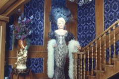 A wax figure of Mae West at Movieland Wax Museum wears an ornate silver dress with white feathered sleeves and a large feathered headpiece, standing on a curved wooden staircase beneath a chandelier in a richly decorated room with blue patterned wallpaper.