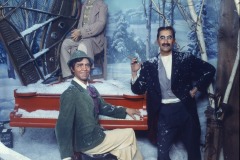 Three Marx Brothers wax figures at Movieland, dressed in old-fashioned winter clothing, pose in a snowy scene with birch trees—one plays a red piano, another sits on top, and the third stands beside them holding a cigar and smiling.