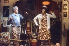 Two older adults strike comedic poses in a vintage living room with patterned wallpaper and retro decor, at Redd Foxx wax figure display at Movieland Wax Museum. The man playfully raises his fists while the woman stands confidently in her floral dress and hat.