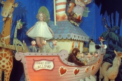 A whimsical display features Shirley Temple in a pink boat labeled Good Ship Lollipop, with a doll-like girl, animals like a giraffe and penguins, all surrounded by candy decorations and a blue tropical backdrop at Movieland Wax Museum.