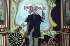 W. C. Fields wax figure at Movieland, Buena Park, California in vintage clothing stands in a carnival-style booth labeled “Professor McGargle’s Rose Tonic”. There are striped curtains and crates of bottles beside him.