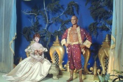 A woman in an elegant, off-shoulder gown sits on a bench beside a figure of Yul Brynner in The King and I at Movieland Wax Museum, standing confidently on a stage with blue drapery, gold décor, and lush greenery in the background.
