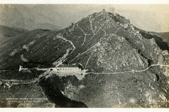 Aerial view of a mountain with winding paths leading to a tavern at its base. The terrain is rocky and rugged, with vegetation scattered across the slope. The image is in black and white, giving it a vintage feel. Text at the bottom identifies the location.
