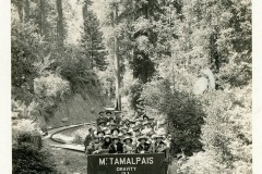 A black and white photo shows a group of people seated in an open railcar marked Mt. Tamalpais Gravity 21 traveling on a narrow track through a dense forest. The track curves gently, bordered by tall trees and foliage.