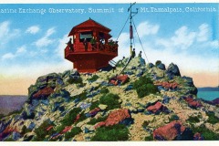 A red-roofed observatory sits on the rocky summit of Mount Tamalpais, surrounded by boulders and sparse greenery. People are standing on the observatory balcony, and a tall antenna extends nearby under a blue sky.