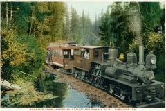 A vintage train travels through a forested area, heading to the summit of Mt. Tamalpais in California. The train consists of a locomotive and passenger car, surrounded by tall trees and lush greenery.