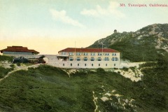 Vintage postcard view of a building on the side of Mt. Tamalpais, California, with a scenic mountainous backdrop. The structure is large, with multiple windows, and a lookout is visible atop the hill. The sky is clear with a few clouds.