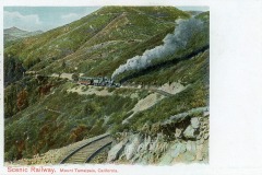 A vintage postcard depicts a train chugging up the scenic, winding tracks of Mount Tamalpais, California, with smoke billowing from the engine. Lush, green hills surround the railway under a partly cloudy sky.