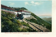 A vintage image shows a mountain train leaving a building at the summit of Mt. Tamalpais, California. The train is on tracks leading down a hill, with a scenic view of hills and a body of water in the distance.