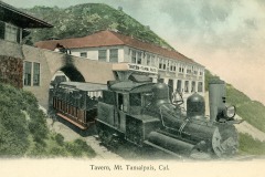 Vintage image of a steam train exiting a tunnel under the Tavern of Tamalpais building, located in Mount Tamalpais, California. The building is nestled against a green hillside with a partly cloudy sky above.