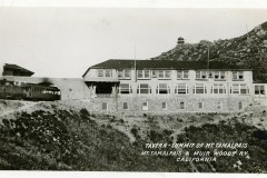 Black and white photo of the Tavern at the Summit of Mt. Tamalpais, with a large building featuring rows of windows. It is surrounded by rocky terrain and a distant elevated lookout. Text: Tavern - Summit of Mt. Tamalpais, Mt. Tamalpais & Muir Woods Ry. California.