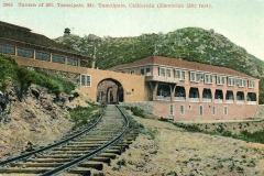 Vintage postcard showing the Tavern of Mount Tamalpais in California. The scene features a building with arches next to railway tracks, set against a backdrop of hills. Text indicates the elevation at 2,592 feet.