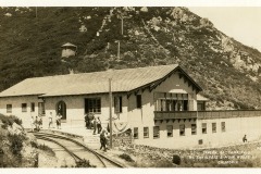 Sepia-toned image of a building near a mountain with Mt. Tamalpais Tavern on it. People stand on a platform next to railway tracks. Rocky terrain and a wooden structure are visible in the background. Text reads Mt. Tamalpais & Muir Woods Ry, California.