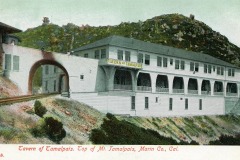 A vintage postcard shows the Tavern of Tamalpais, a large white two-story building with arches and a sign. Its situated on a hillside with a railroad track leading into a tunnel. The background features a mountain and greenery.