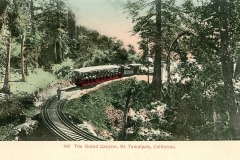 A vintage postcard image depicts a red train traveling on curved tracks through a lush forested area on the side of a mountain. The scene is labeled The Grand Canyon, Mt Tamalpais, California.
