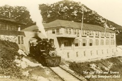 A vintage steam train emerges from a tunnel beneath a large wooden building, labeled The Old Train and Tavern atop Mt. Tamalpais, Calif. Smoke billows from the train. The landscape is rocky with hills in the background.