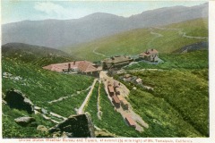 A vintage image showing buildings at the summit of Mt. Tamalpais, California. The structures, including the United States Weather Bureau and Tavern, are surrounded by green hills and winding paths under a clear sky.