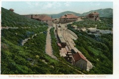 A vintage postcard shows the United States Weather Bureau and the Tavern at Summit on Mount Tamalpais, California. The scene includes buildings nestled in lush greenery with a mountain backdrop and a narrow path leading through the landscape.