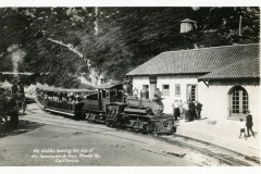 A vintage steam train at a station on the Mt. Tamalpais & Muir Woods Railway in California. Several people stand on the platform next to the train. The background features hills with trees and vegetation. The image is in black and white.