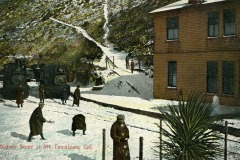 A vintage winter scene at Mt. Tamalpais, California, featuring people walking through snow near two train engines. A brown wooden building stands on the right, and a snowy hill with a fence is in the background.