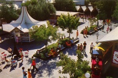 A vibrant outdoor amusement park scene with people walking, sitting, and enjoying various attractions. Theres a carousel, food vendors under a tent, trees, and benches. Bright clothing and shadows suggest a sunny day.