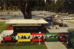 A miniature train with colorful carriages is passing by Nut Tree Airport. Several small airplanes are parked in the background. People are walking nearby. A sign reads Nut Tree Airport. The scene is sunny with trees in the background.