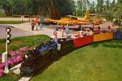A small train with passengers rides along a grass field near a parked yellow airplane. Nearby, people stand, and children watch. The scene is surrounded by lush trees and a bright sky, creating a cheerful, vintage amusement park atmosphere.