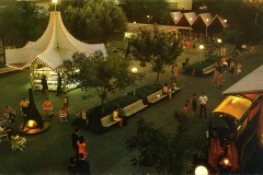 An aerial view of a lively amusement park at dusk, featuring a lit-up carousel-style booth, numerous benches, and trees. People are strolling, seated, or gathered in small groups, while a train ride is visible in the foreground.