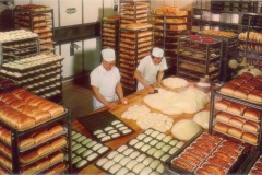 Bakery with two bakers in white uniforms working. Large trays of bread and rolls are stacked around them. The bakers are shaping dough on a central wooden table, surrounded by ovens and racks filled with baked goods.