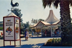 A vintage photo of a plaza with a white tent-like structure. A sign with a rooster on top reads Dining Rooms and displays icons of food. People are walking around, and a palm tree is on the right.