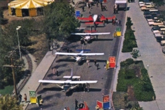 Aerial view of a park with several small airplanes lined up on a paved area. People walk around nearby. On the left, a circular structure with a yellow and white-striped roof is visible. Trees and parked cars surround the area.
