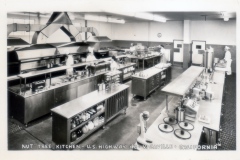 Black and white photo of a large, industrial kitchen labeled Nut Tree Kitchen - U.S. Highway 40 - Vacaville - California. It features chefs at workstations, stainless steel equipment, and shelves stocked with jars and utensils.