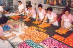 Workers in a candy factory wear pink uniforms and white aprons while sorting colorful, striped candies. They are standing around a long table, organizing candies with vibrant swirling patterns in orange, red, blue, and purple.