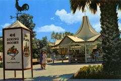 A vintage outdoor scene with a dining rooms sign featuring playful illustrations. People stroll around a plaza with a tented pavilion. A weathervane with a rooster sits above. The area is surrounded by trees and a palm tree.