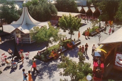 A vibrant amusement park with various attractions and people walking around. Theres a carousel-like structure, trees lining the paths, red chairs scattered, and visitors sitting on benches. The scene is lively and colorful, with bright clothing visible.