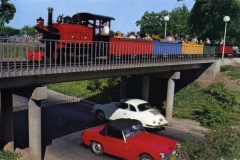 A small train painted in red, yellow, and blue with passengers sits on a bridge above a road. Below, two cars, one white and one red, are driving. Trees and street lamps surround the area.