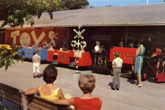 A mini train with colorful carriages, numbered 5, is at an amusement park station. A train conductor stands nearby. Two women and a child are seated on a bench in the foreground, while others stand and observe. Toy sign and railroad crossing visible.