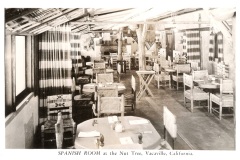 Black and white photo of the Spanish Room at the Nut Tree in Vacaville, California. The room features rustic wooden tables and chairs, striped curtains, and decorative items on the walls. Tables are neatly set with tableware.