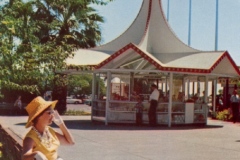 A woman in a yellow hat and dress sits on a bench, shielding her eyes from the sun. Behind her is a pavilion with a red-and-white roof. Three flags, including the American flag, are visible against the clear blue sky in the background.