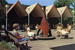Vintage scene of people sitting around a conical outdoor fireplace at a rest area. The setting includes angled roof structures, trees, and people dressed in mid-century attire, some holding cups. The atmosphere is relaxed and social.