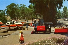 A small train with the word RAILROAD on the side is carrying passengers by a nearby parked light airplane. Trees line the background, and people, including children, are walking around in bright sunlight.