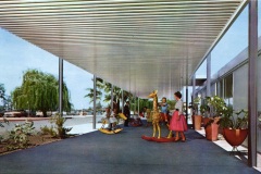 Children playing with colorful rocking toys under a modernist outdoor canopy. A woman stands nearby, and large planters with greenery line the walkway. Trees and a clear sky are visible in the background.