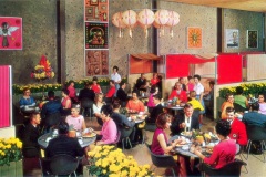 A vibrant restaurant scene from the mid-20th century, featuring people dining at round tables with a colorful decor. The room is filled with yellow flowers and large, artistic wall hangings, creating a lively and cheerful atmosphere.
