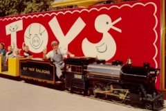 Children and a conductor ride a small train marked Nut Tree Railroad in front of a bright red sign with the word TOYS and playful illustrations, including a pig and a duck.