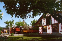 A colorful train with Santa Fe & Disneyland branding is stationed near a glass building adorned with large alphabet blocks. Trees provide shade, and a few people are visible in the background under a clear blue sky.