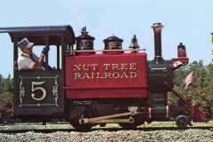 A small, vintage steam train labeled Nut Tree Railroad with a large number 5 on its side. A person in a conductors uniform is seated in the cabin. The train features an American flag and ornate details.