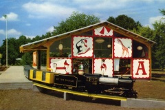 A small, colorful train labeled Nut Tree Railroad is stationed on tracks in front of a building. The building has large cartoon images of animals and a circus theme. Trees are in the background, and the scene is bright and sunny.