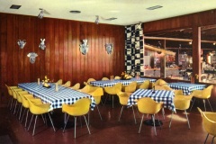 A dining room with wooden walls, decorated with wall-mounted animal heads, features tables covered with blue and white checkered cloths. Yellow chairs surround the tables, and a patterned wall divides the room from a busy background area.