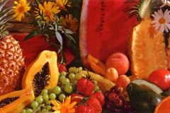 A colorful assortment of fruits including a sliced watermelon, pineapple, papaya, strawberries, grapes, and bananas, surrounded by sunflowers and daisies, arranged against a wooden background.